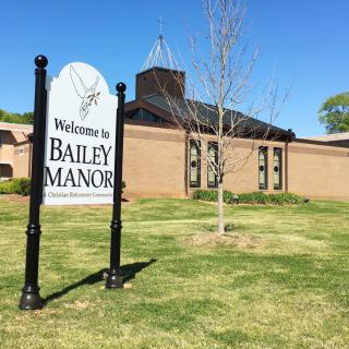 A sign in front of a building reads 'Welcome to Bailey Manor' on a sunny day.