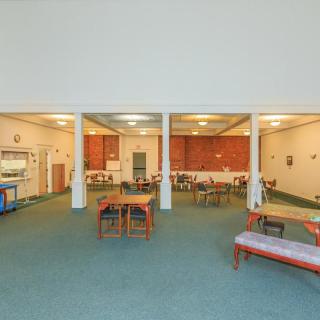 A spacious dining area with multiple tables and chairs, featuring large windows and simple furnishings.