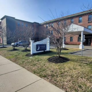A sunny view of a building with a sign on the front lawn and parked cars nearby.