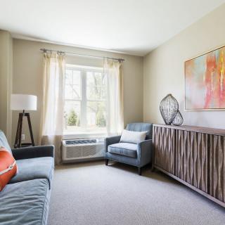 A cozy living room with modern furnishings, including a sofa and armchair, accented by a large window and colorful artwork.