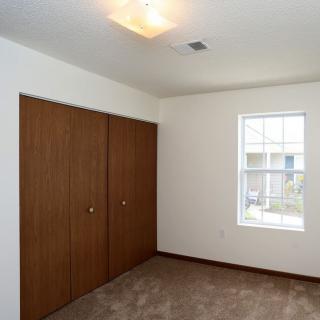 An empty bedroom with a carpeted floor, featuring a window and wooden closet doors.
