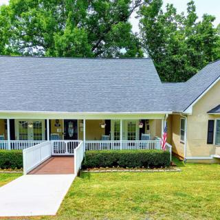 A charming single-story house with a front porch and accessibility ramp, surrounded by a well-kept lawn and trees.