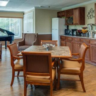 A cozy dining area with a kitchen in the background, featuring a table with chairs and a piano by the window.