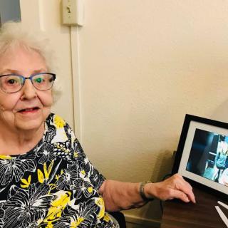 An elderly woman sitting by a desk, smiling and pointing to a photo in a frame.
