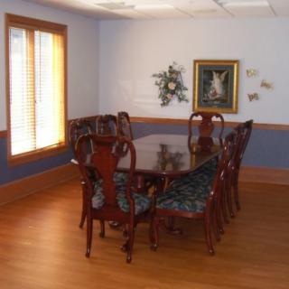 A dining room featuring a wooden table surrounded by chairs, with a painting on the wall and natural light coming through a window.