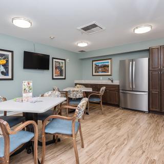 A cozy dining area with tables, chairs, and a refrigerator in the corner, adorned with artwork on light blue walls.