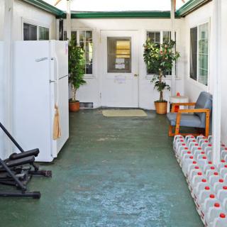 A covered patio with a refrigerator, seating area, and exercise equipment, lined with water bottles and potted plants.
