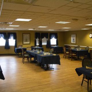 An empty dining room with tables and chairs set up, featuring wooden floors and decorated windows.