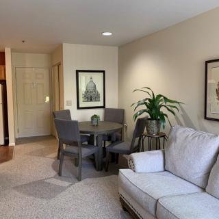 A cozy living room with a small dining area, featuring a sofa, chairs, a round table, indoor plant, and framed artwork on the walls.