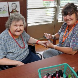 A caregiver is happily painting an elderly woman's nails at a table indoors.