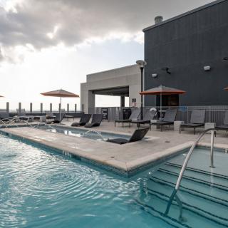 A modern rooftop pool with sun loungers and umbrellas overlooks an urban skyline under a cloudy sky.