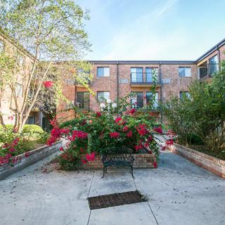 A scenic courtyard featuring a brick apartment building, lush greenery, and vibrant flowers with a central bench.