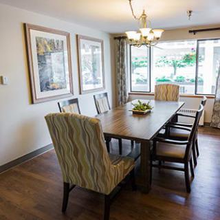 A cozy dining room with a wooden table, surrounded by chairs, and decorated with artwork on the walls. Large windows bring in natural light.