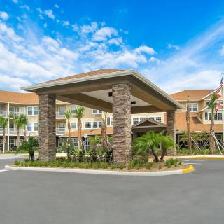 A modern building entrance with a covered driveway and surrounding palm trees under a partly cloudy sky.