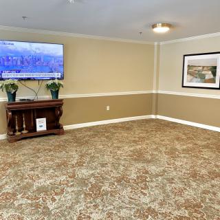 An empty living room with a carpeted floor, featuring a wall-mounted television, a painting, and some furniture.