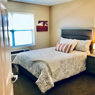 A cozy bedroom with a neatly made bed, featuring patterned bedding and decorative pillows. The room is softly lit by a bedside lamp, with a window allowing natural light to brighten the space.