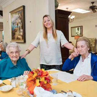 A caregiver stands between two elderly women seated at a table with autumn decorations.