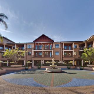 A sunny courtyard featuring a brick building with balconies and a central fountain, surrounded by trees.