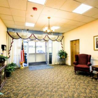 A warmly lit lobby with a carpeted floor, featuring armchairs, potted plants, and an American flag near the entrance.