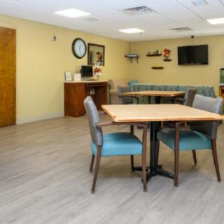 A clean and organized dining or community space with tables, chairs, a television, and a wall clock.