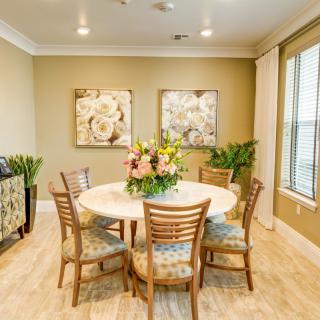 A cozy dining room with a round table surrounded by wooden chairs, featuring a floral centerpiece and decorative art on the walls.