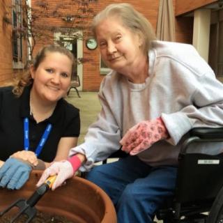 An elderly woman in a wheelchair enjoys gardening with the assistance of a caregiver.