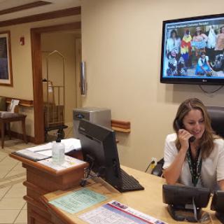 A receptionist attending to a call at the front desk of an office, with a monitor displaying a group photo.