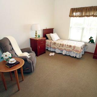 A cozy bedroom featuring a single bed with a bedside table and lamp, an armchair with a side table, and a window with curtains.