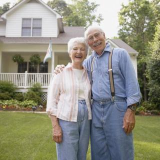 An elderly couple standing in front of their house, smiling and enjoying a sunny day.