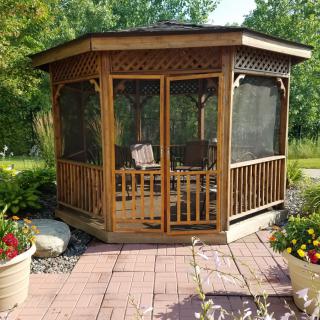 A wooden, screened gazebo sits on a tiled patio amidst lush garden surroundings.