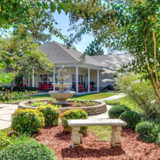 A beautifully landscaped garden with a central fountain and stone bench, surrounded by lush greenery and leading up to a cozy house.