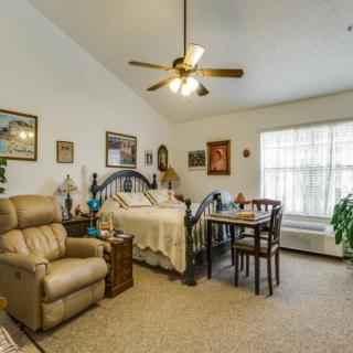 A cozy living space with a bed, armchair, table, and decorative elements. A ceiling fan hangs from the ceiling, and sunlight streams through the large window.