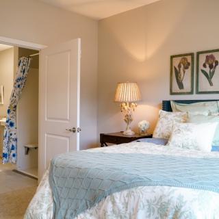A cozy bedroom with elegant decor, featuring a neatly made bed, stylish lamp, and floral artwork.