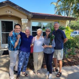 A group of people smiling and posing together outside a house on a sunny day.