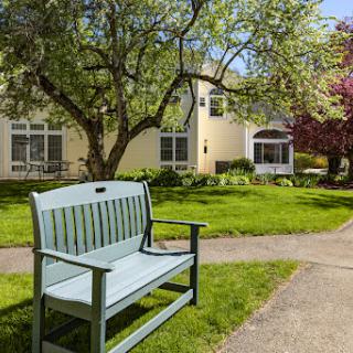A serene outdoor space featuring a green bench surrounded by lush grass and trees, with a cozy building in the background.