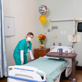 A nurse in a hospital room prepares a bed with flowers and a balloon nearby.