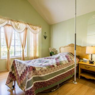 A cozy bedroom with a quilt-covered bed, large windows with curtains, and a bedside lamp on a wooden floor.