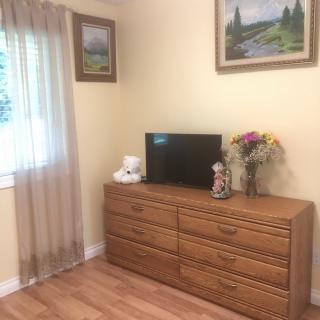A cozy bedroom corner featuring a wooden dresser topped with a television, decorative items, and flowers, alongside a window with sheer curtains and framed paintings on the wall.