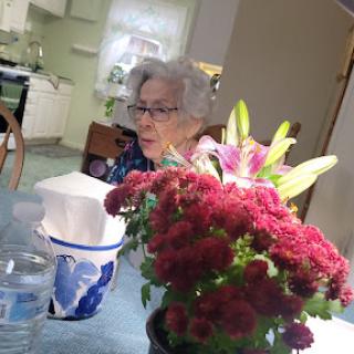 An elderly woman seated at a table surrounded by vibrant flowers in a cozy kitchen setting.