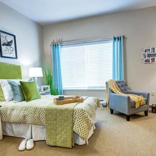 A cozy and modern bedroom with elegant decor, featuring a bed with green accents, an armchair by the window, and soft natural lighting.