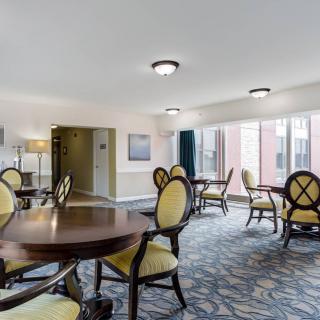 A well-lit room with round tables and cushioned chairs, featuring large windows and patterned carpet.