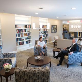 Two people having a conversation in a library setting with bookshelves and comfortable seating.