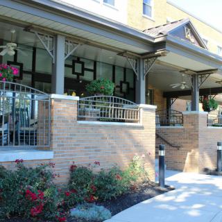 The image shows the entrance of a building with a porch and brick walls, surrounded by landscaping and flowers.