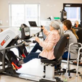 An elderly person engaging in a workout session using specialized exercise equipment with assistance from a caregiver.