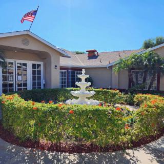 A house with a beautifully landscaped garden featuring a central fountain, adorned with an American flag and surrounded by palm trees on a sunny day.