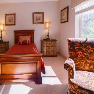 A cozy bedroom featuring classic wooden furniture, including a bed and bedside tables with lamps. An armchair sits by the window, allowing natural light to fill the space.
