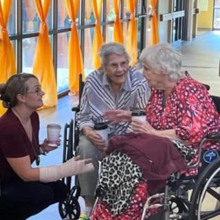 Three women engage in a warm conversation, two seated in wheelchairs and one kneeling beside them, sharing a moment of companionship indoors.