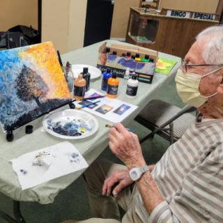 An elderly person wearing a mask is engaged in painting a colorful canvas indoors, surrounded by art supplies.