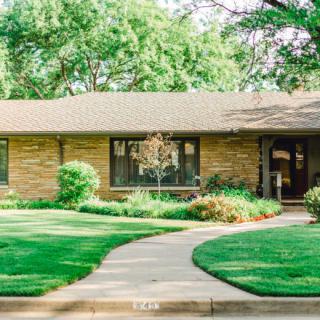 A brick house surrounded by a well-manicured lawn and trees, with a curved concrete pathway leading to the entrance.