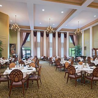A spacious and elegantly decorated dining room with multiple tables and chairs, featuring tall windows and chandeliers.